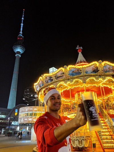 a man standing in front of a carousel at alexanderplatz christmas market in berlin with the tv tower in the background