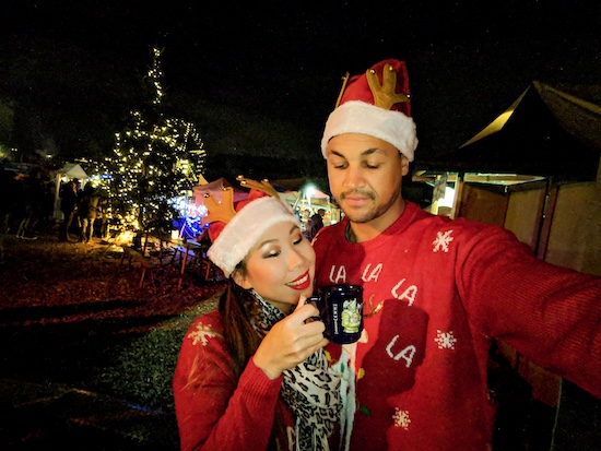 a couple wearing red xmas jerseys looking at a mug of gluhwein with food stalls in the background