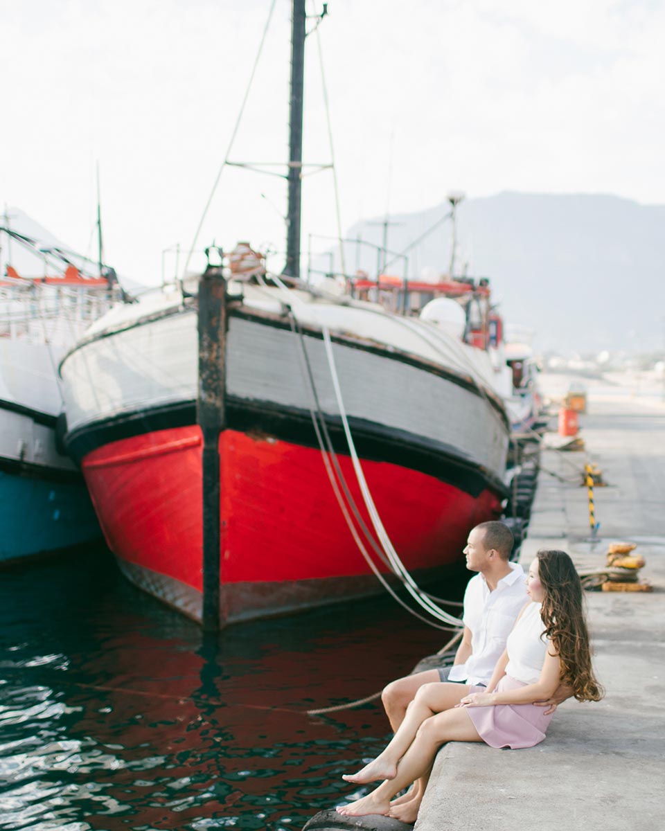 a couple sitting on the docks at hout bay harbour with a red and white fishing boat in the background