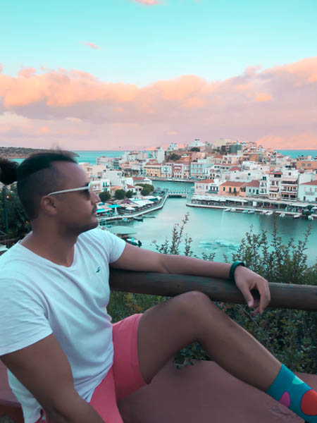 a man sitting on a ledge at a viewpoint overlooking agios nikolaos in crete