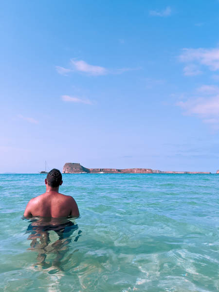 a man standing in crystal clear waters at Balos Beach with Gramvousa Island in the backdrop