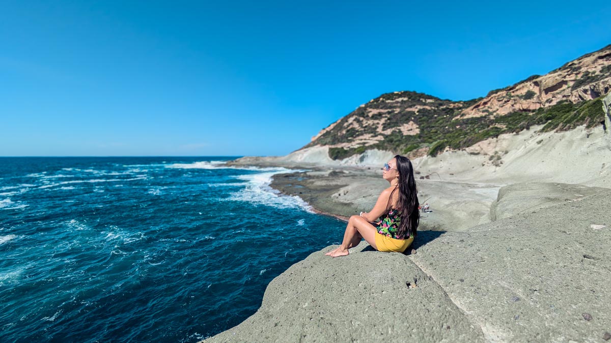 a woman sits on the edge of a gray rocky beach with the sea and hills in the background near the town bosa