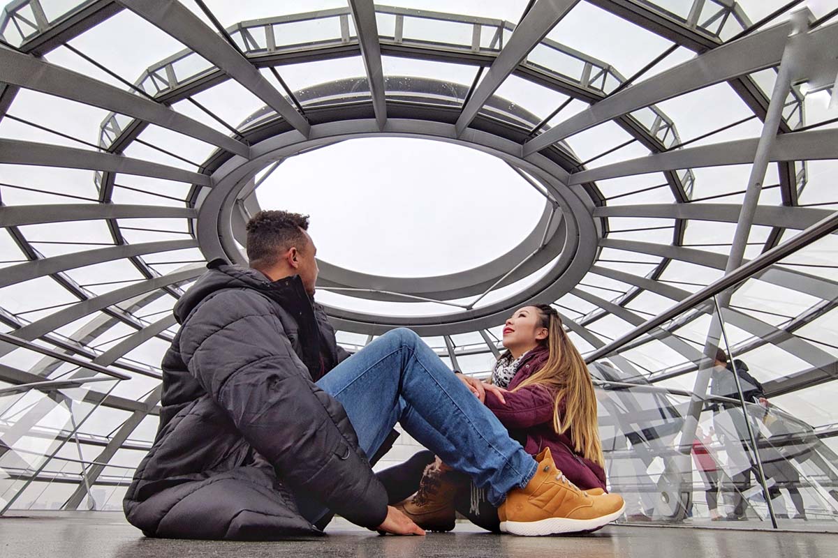 a man and a woman seated on the floor inside the Dome of the Reichstag Building gazing upwards while admiring the architecture