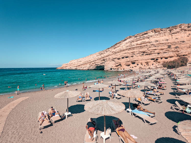 a view of matala beach with beach umbrellas and sunbeds on white sand and matala caves in the background