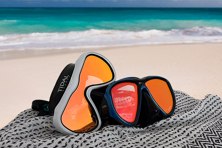 two anti fog snorkel and scuba masks lying on a sarong on a beach with white sand and turquoise blue waters