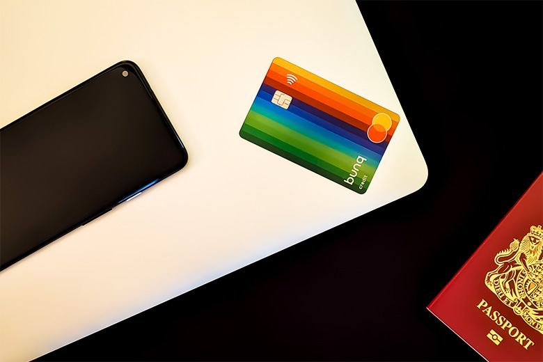 the iconic rainbow bunq bank card placed next to a black mobile device and both placed on top of a silver laptop which is lying next to a maroon passport