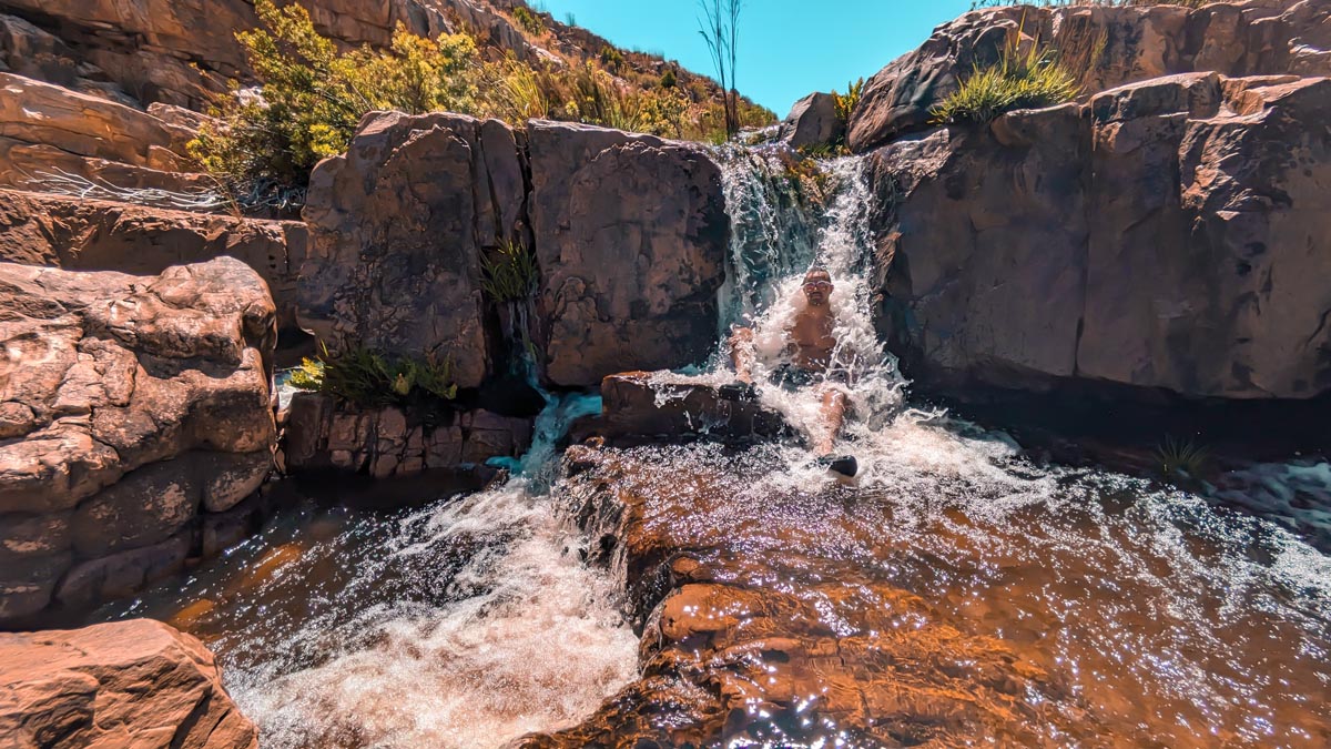 a man sitting under a waterfall at maalgat rock pool in cederberg south africa 