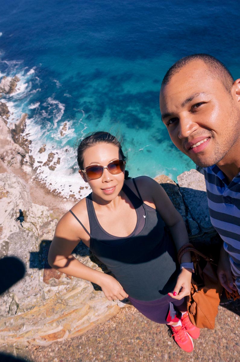 a couple taking a selfie with a view of the blue ocean below at cape point national park