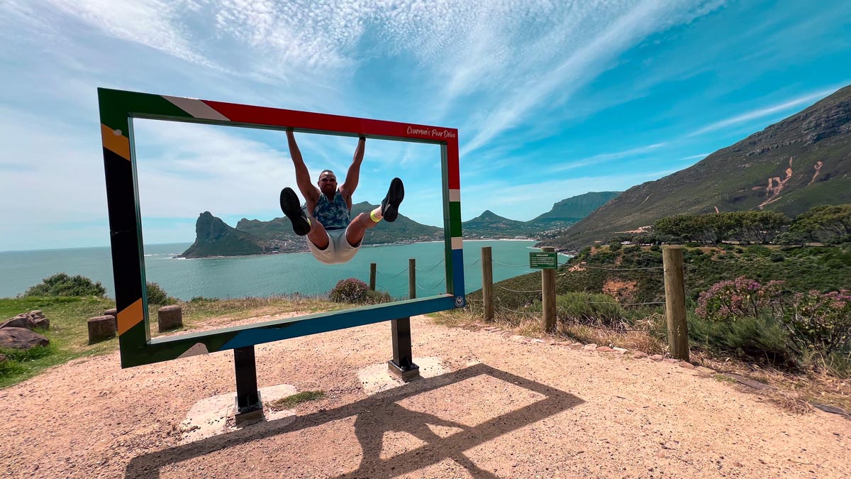 a man taking a photo hanging on the iconic photo frames along chapman's peak drive with the mountain and ocean in the background 