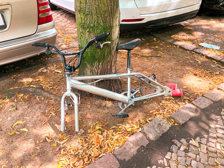 a vandalised bicycle lying against a tree with its wheels stolen