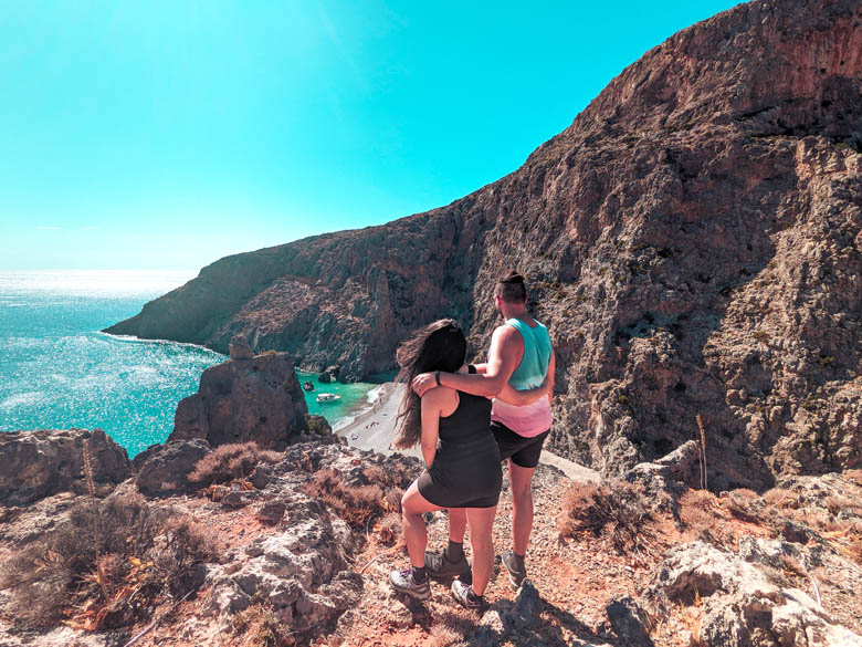 a couple standing on the edge of a cliff looking down at agiofarago beach