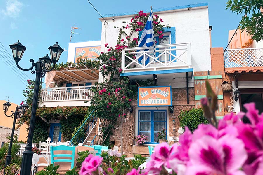 a picturesque blue and white building in crete greece with tables and chairs outside and flowers growing on the wall