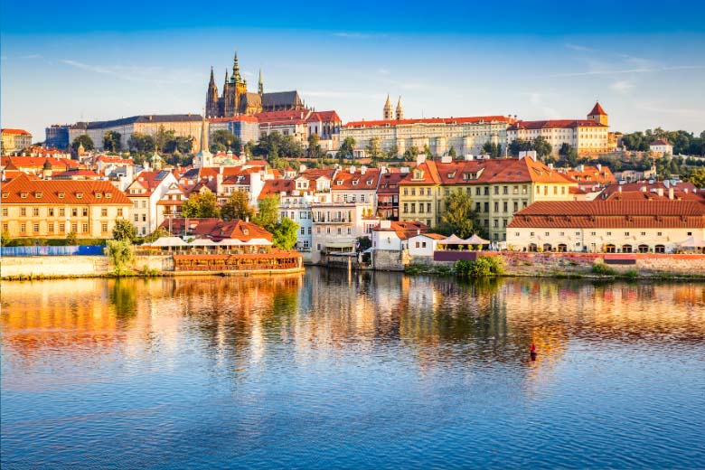 a long shot of Prague Castle positioned above the city littered with red roof buildings in front of the Vltava River taken from Charles Bridge