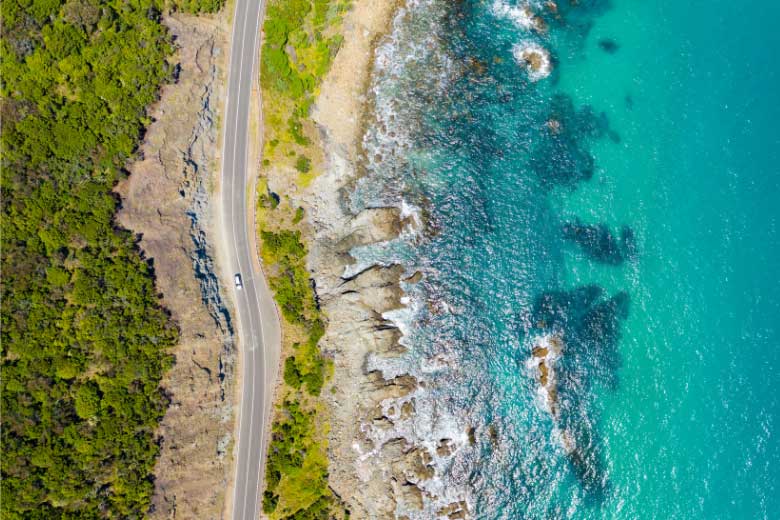 a drone shot from above of a car driving on a road in crete along the ocean on one side and trees on the other