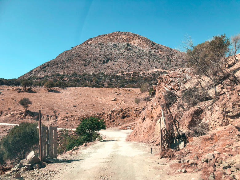 an unpaved, dirt road with a mountain in the backdrop leading to agiofarago beach in crete, greece