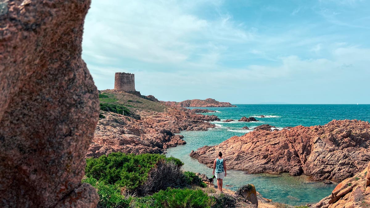 a man and dog hiking along the coastline near isola rossa with a tower and the seas in the background