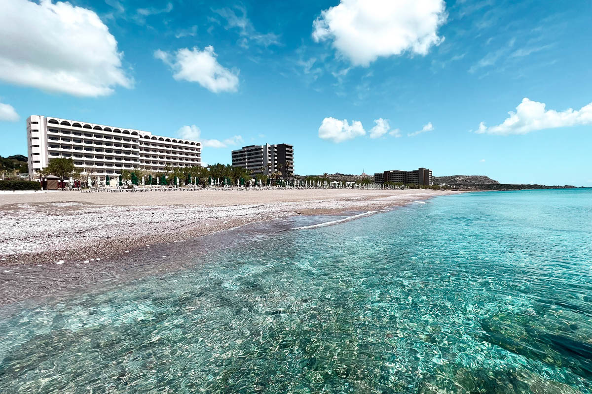 a view of faliraki beach with crystal clear turquoise blue waters and a hotel building in the background