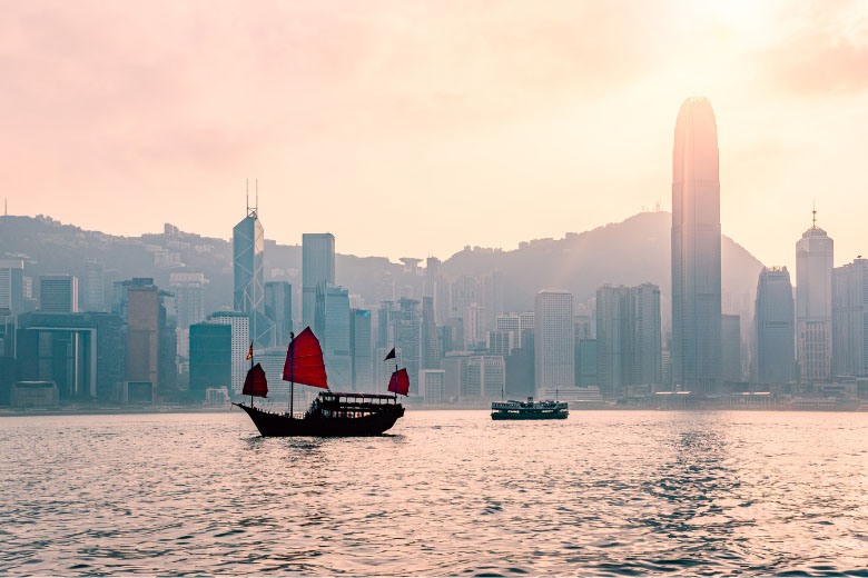 a Junk boat with three red sails sailing ahead of a ferry across the waters of Victoria Bay with the buildings of Hong Kong Island in the background
