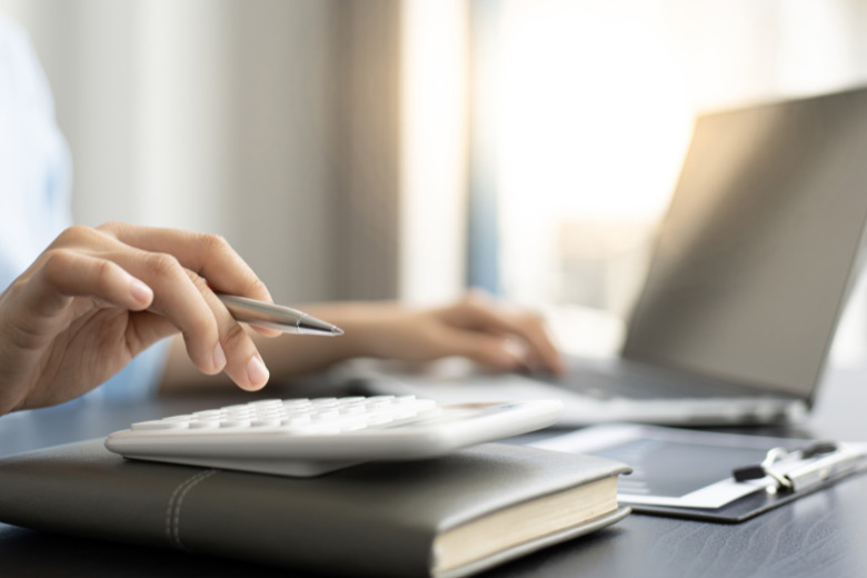 a person seated at a desk inputting into a calculator while typing on a laptop