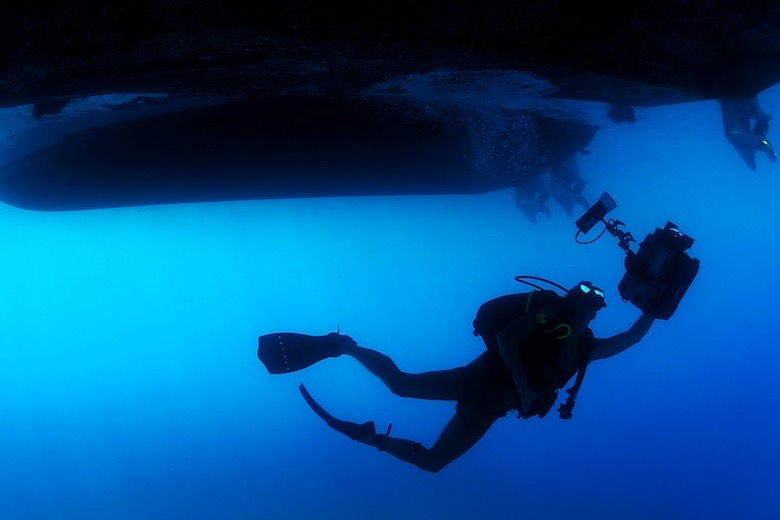 a man scuba diving with underwater camera equipment in hand ascending to two boats waiting on the surface