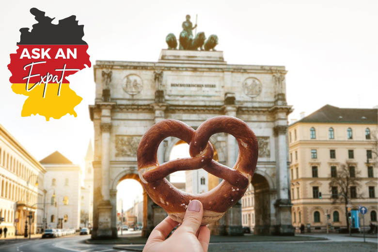 a person holding a pretzel in front of a monument in munich germany and the icon of a german flag with the text ask an expat on the top left hand corner