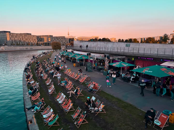 people sitting in sun loungers next to the canal in the city centre of berlin enjoying drinks and the sunset