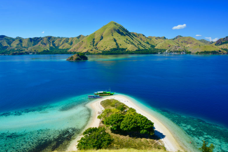 an aerial view from on top of a hill overlooking the tip of an island with boats anchored on the shores of a white sandy beach surrounded by crystal clear turquoise blue water with a mountainous mainland covered in green vegetation in the far distance
