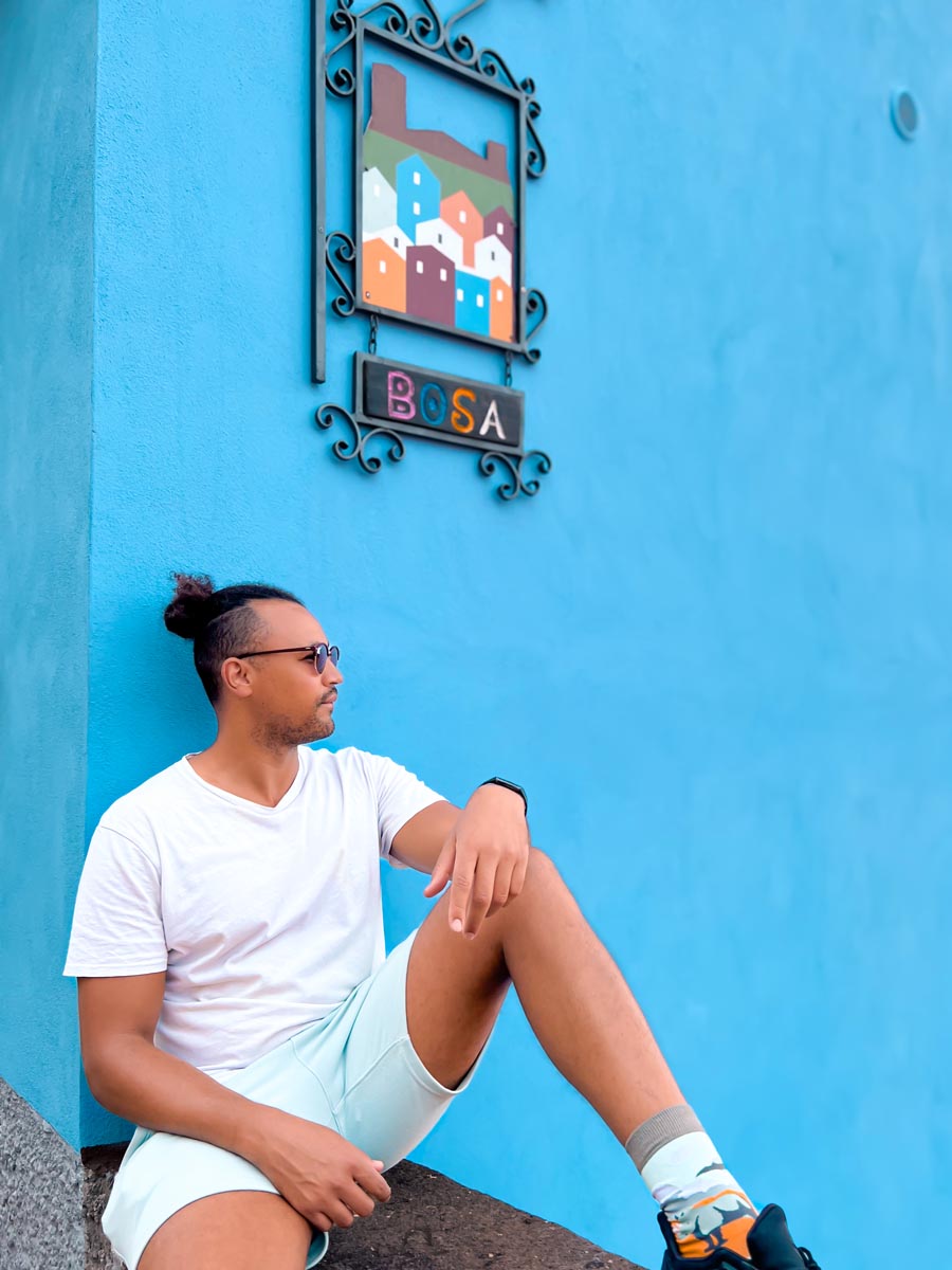 a man sitting on a ledge against a blue wall looking into the distance with the sign bosa above him in north west sardinia italy
