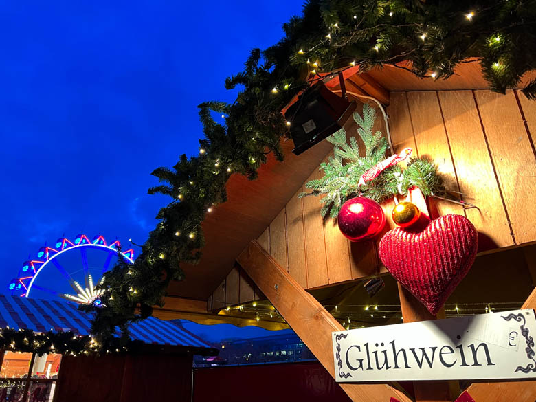 a xmas market stall in germany with a gluhwein sign and a giant ferris wheel in the backdrop