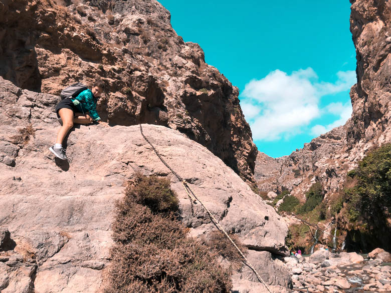 a woman scrambling on all fours climbing large rocks next to the kourtaliotiko river