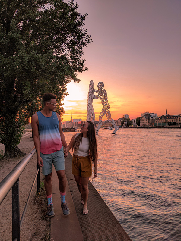 a couple walking hand in hand along the Spree River at sunset, with the Molecule Man sculpture in the background, showcasing the perfect balance of Berlin's vibrant city life and tranquil natural beauty