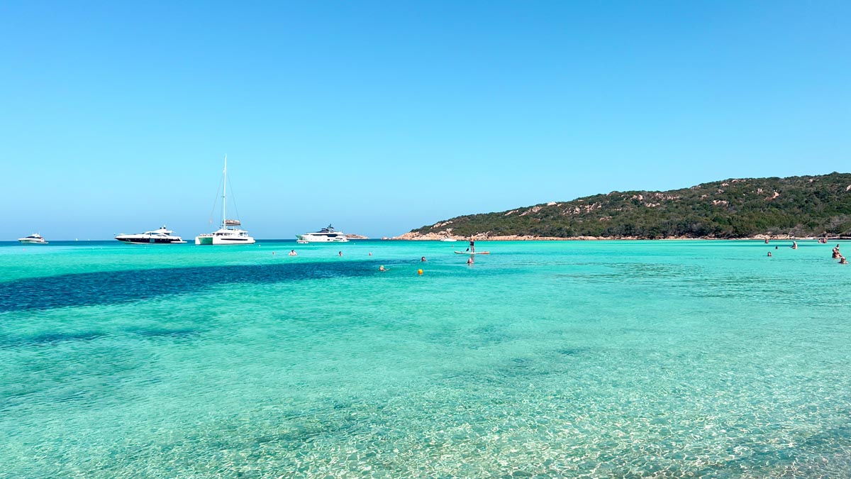 a beach with crystal clear turquoise blue waters and yachts floating in the distance in north sardinia on a blue sunny day