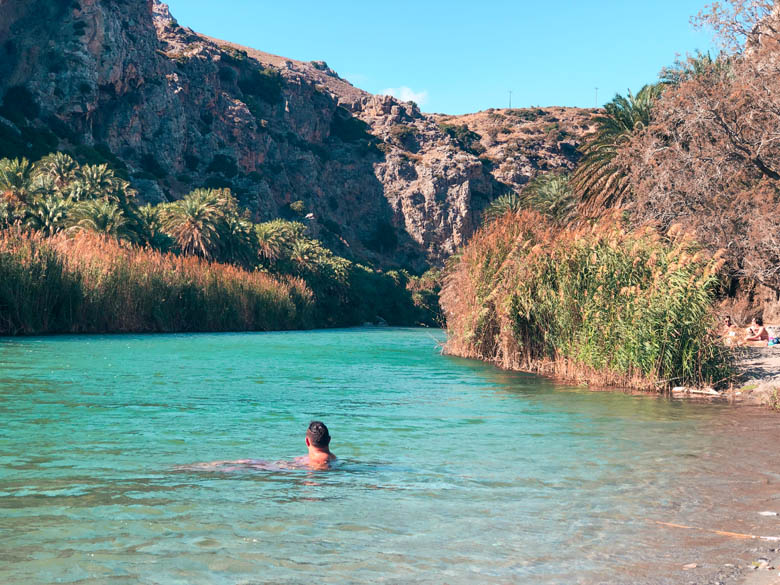 a man lying in water at preveli beach with palm tree forest in the background