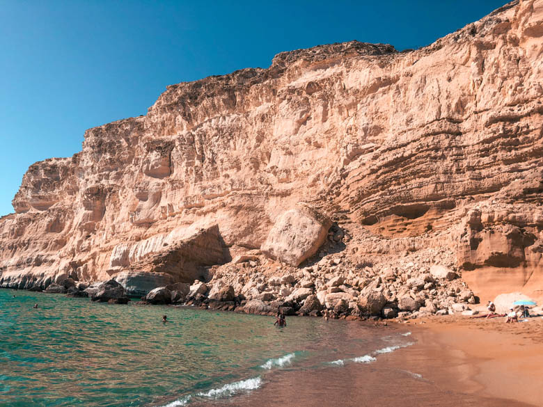 a reddish orange sandy beach with turquoise blue waters at the end of the red beach trail in crete greece