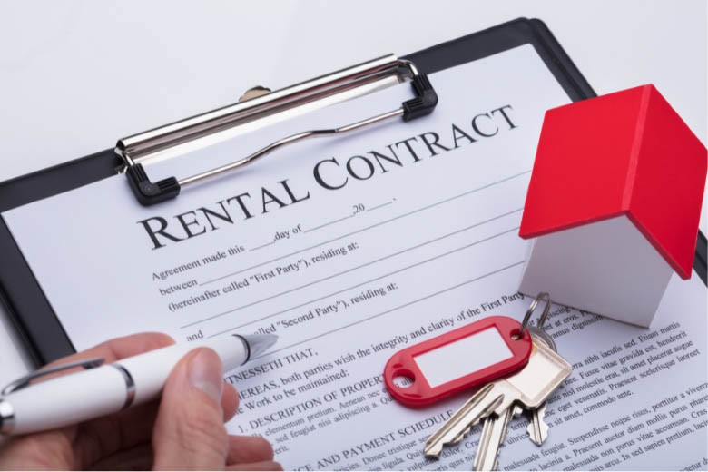 a hand holding a pen over a rental contract alongside a set of keys and a small house model, symbolizing the signing of a lease agreement