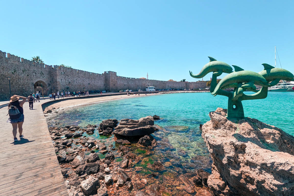 a traveler walking along the promenade near old town rhodes with a view of the sea and dolphin sculpture