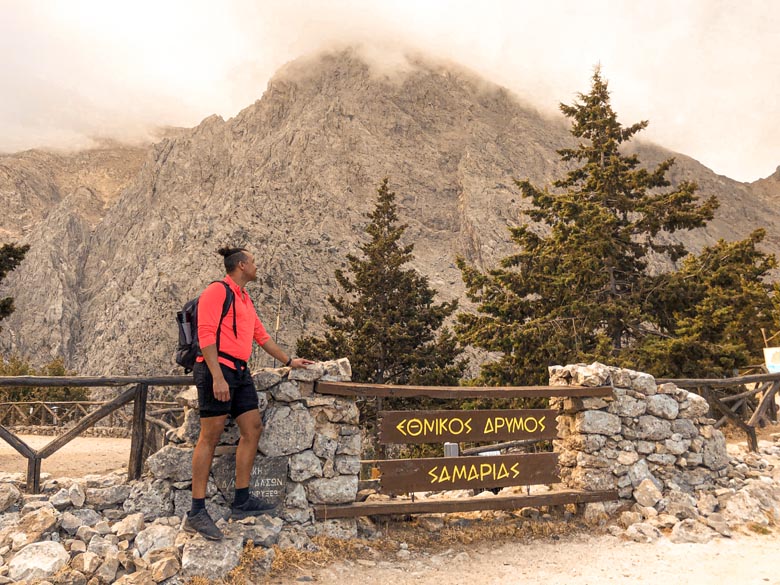 a man standing near the entrance to samaria gorge national park with the white mountains of crete in the background