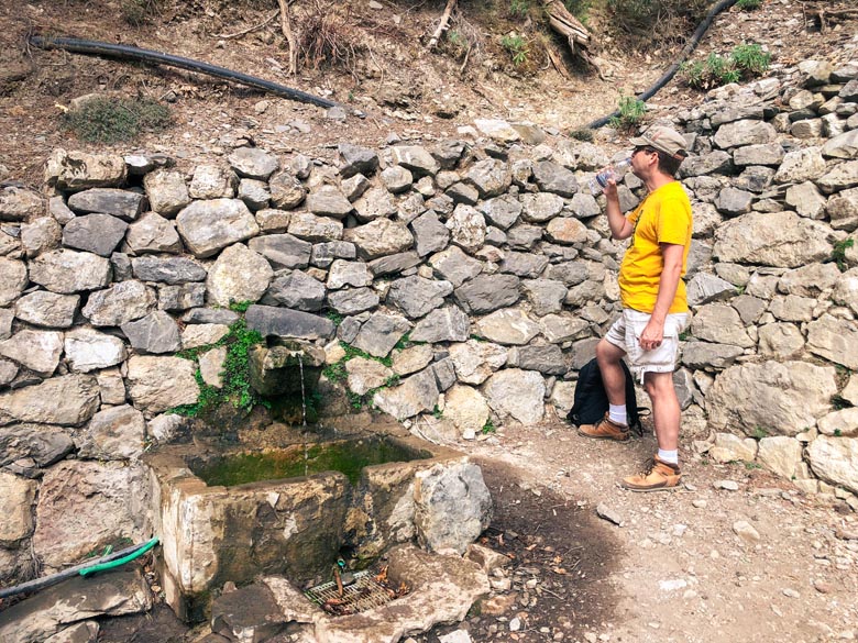 a man drinking water next to a fountain on a hike in crete