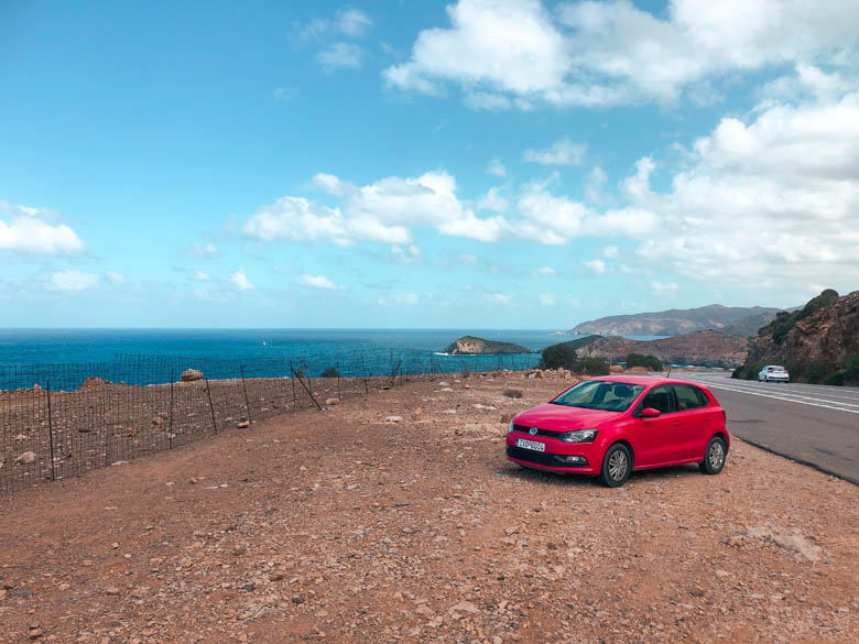 a red vw car rental parked on the side of the road in crete with a backdrop of the ocean