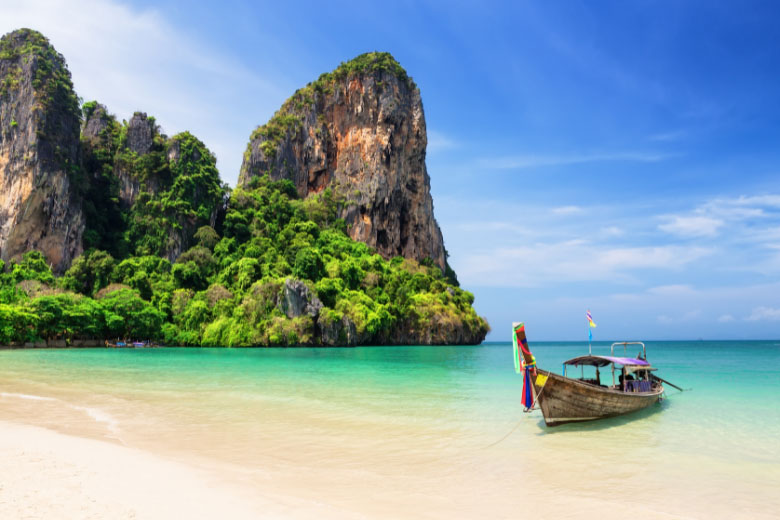 a long-tail boat anchored on the shores of Railay Beach with limestone cliffs covered in lush green vegetation in the background