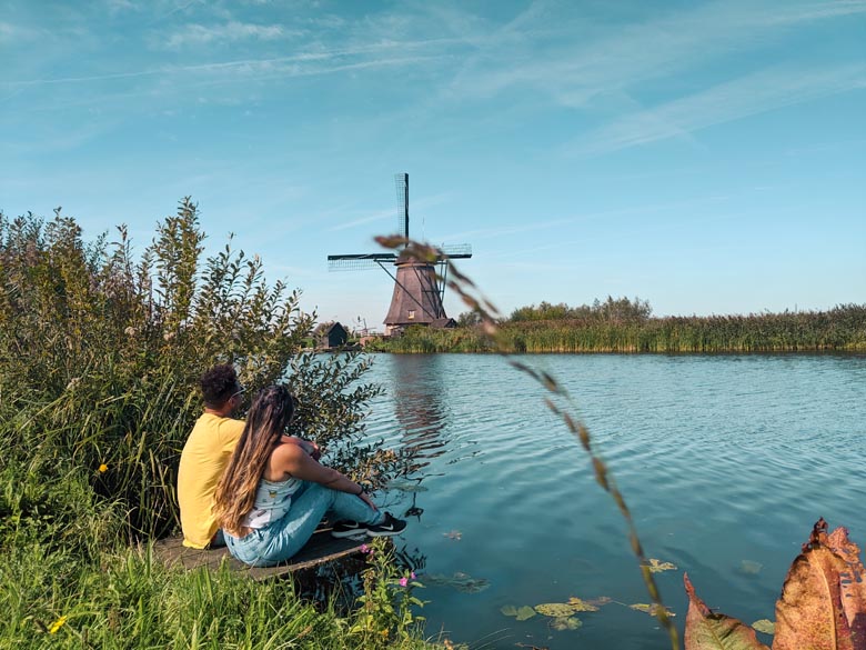 best photography spot in the netherlands with a couple sitting next to the river and a traditional windmill in the background