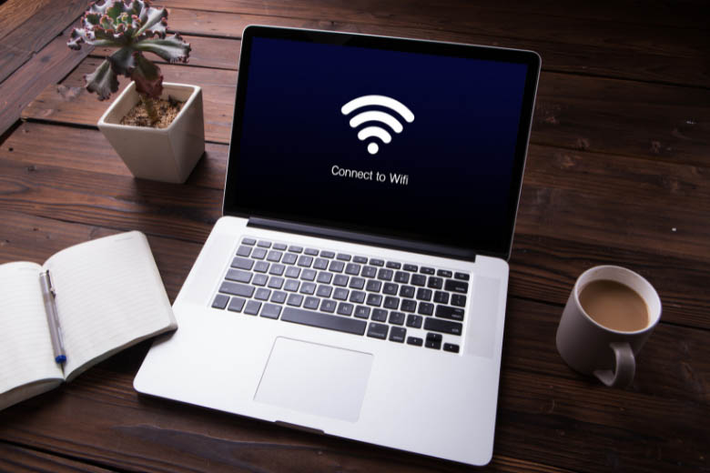 a laptop connecting to wifi on a dark wood table lying next to a notebook and cup of coffee