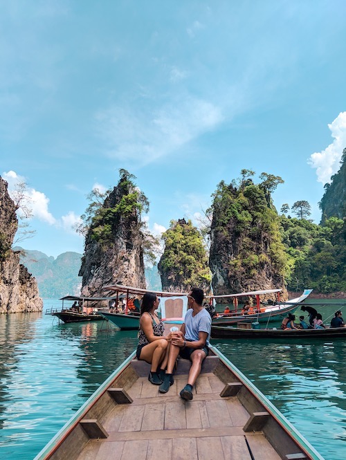 a woman and a man sitting on the bow of a long-tail boat looking back over their shoulders while admiring limestone cliff islands in Thailand