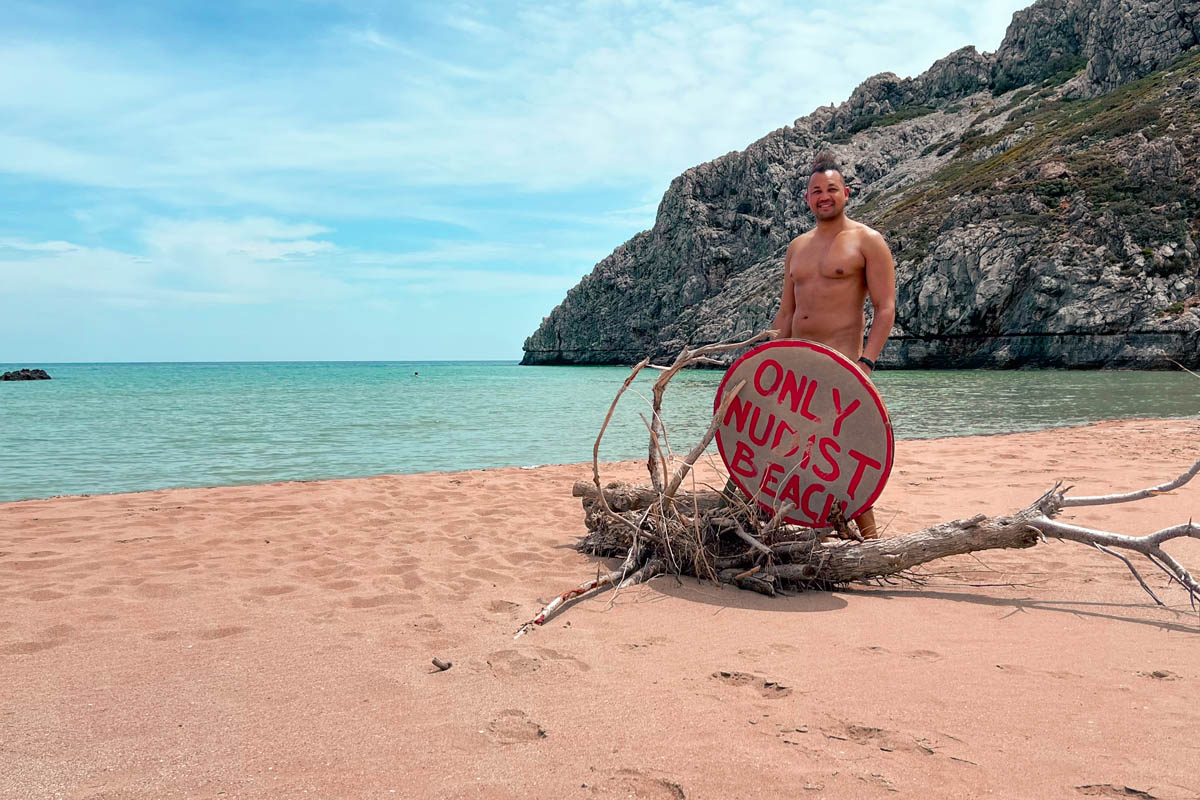 a man standing behind sign that says nudist beach with a view of the sea in the background