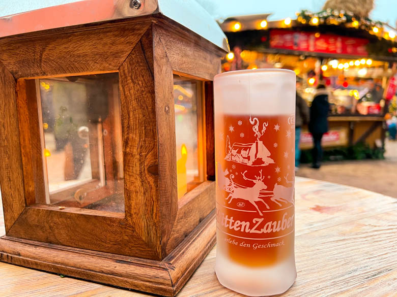 a mug of rose gluhwein next to a candle in a wooden box with colorful xmas stalls in the background