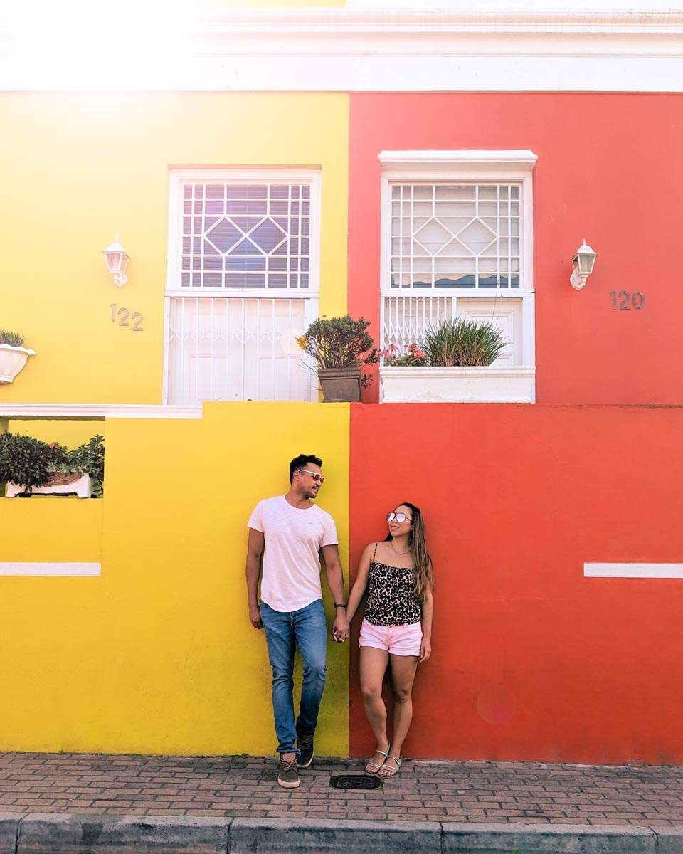 a couple standing against a bright yellow and orange painted buildings in bo-kaap city centre of cape town at the intersection of wale street and chiappini street a famous instagram photo spot