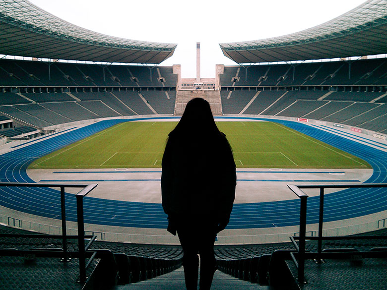 a silhouette of a woman standing on the spectator stands with the view of the berlin olympic stadium in the back